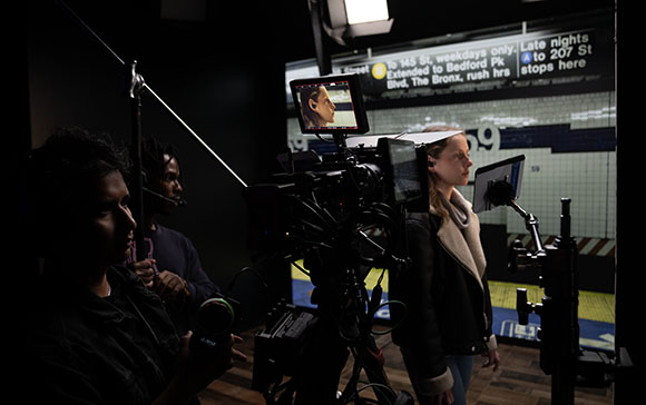 A film crew operates a professional camera in a dark studio with a large digital screen behind them.