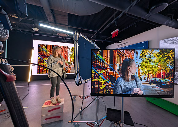 A woman stands in a studio next to a large monitor displaying a vibrant street scene with autumn trees.