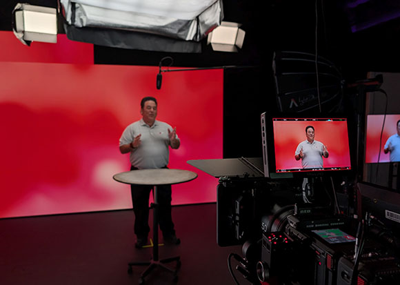 A man stands behind a high table in a studio with a red background, viewed through a professional camera monitor.