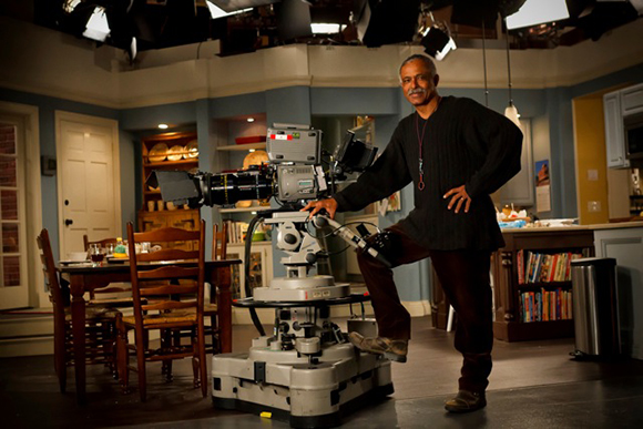 A man in black clothing stands with one foot on a large professional studio camera in a kitchen set.