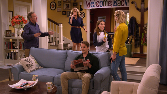 Five people in a living room; a man sits on a blue sofa while others stand and talk in the background.