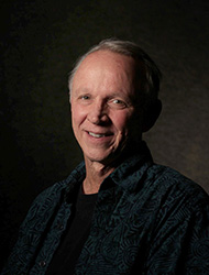 A smiling older man with short grey hair wearing a dark patterned button-down shirt.