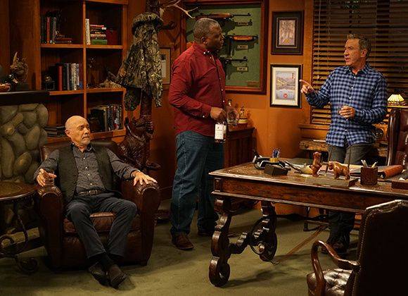 A man in a red shirt stands between a seated man in a leather chair and a man in a blue plaid shirt.