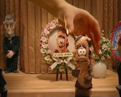 A hand reaches toward a small, carved wooden figure in front of a floral memorial portrait.
