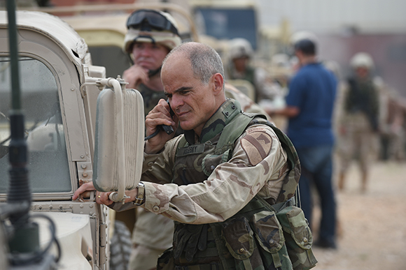 A man in camouflage fatigues talks on a mobile phone while standing beside a military vehicle.