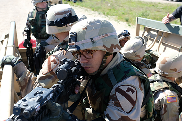 Soldiers in desert camouflage helmets and gear riding in a military vehicle, one aiming a rifle.