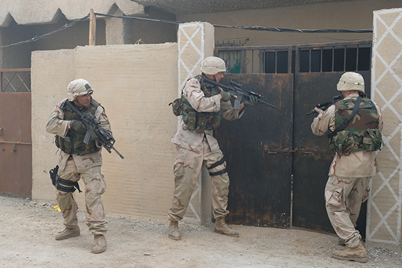 Three soldiers in desert camouflage gear approach a dark metal gate in a sandy environment.