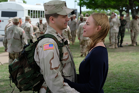 A soldier in desert camouflage and a woman in a dark blue shirt look at each other closely outdoors.