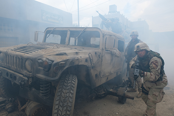 Soldiers stand near a tan military vehicle amidst thick smoke and dust.