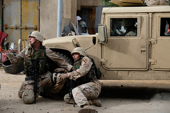 Two soldiers in desert camouflage crouch behind a tire near a tan Humvee on a dusty street.