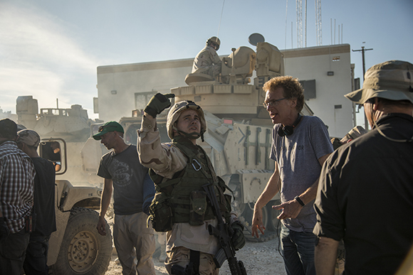 A soldier in desert gear gestures upward while a man in a blue shirt talks to him near military vehicles.