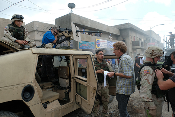 Soldiers and men in civilian clothes stand near a tan Humvee with a mounted machine gun in a dusty urban area.