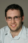 A man with short dark hair and glasses wearing a light-colored button-down shirt.