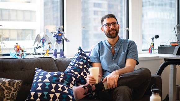 A man with glasses and a beard sits on a dark sofa holding a white cup near a window with toys.