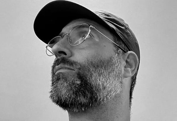 Black and white close-up of a man with a beard, wearing glasses and a baseball cap, looking upward.