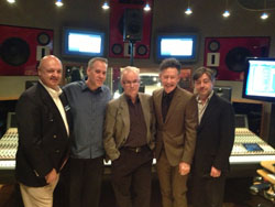 Five men in suits and casual wear stand in a recording studio in front of a mixing board and monitors.