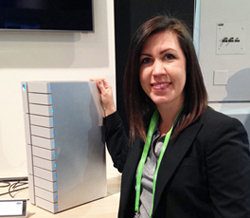 A woman in a black blazer smiling while standing next to a tall, silver server rack with blue lights.