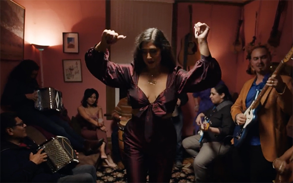 A woman in a purple satin outfit dances with raised arms in a room with musicians playing accordion and guitar.