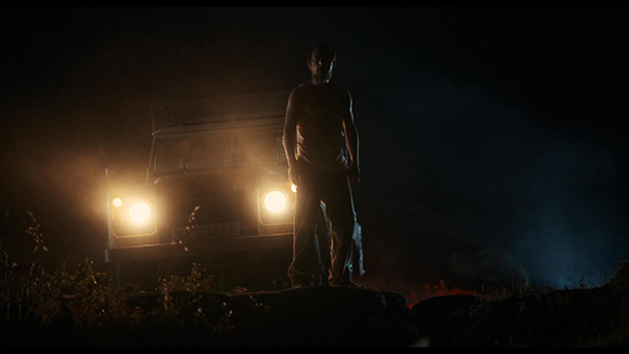 A man stands in silhouette in front of a vehicle with bright headlights on at night.