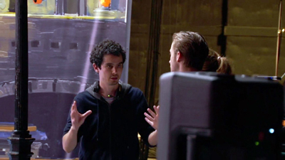 A man in a dark hoodie gestures with both hands while speaking to someone off-camera.