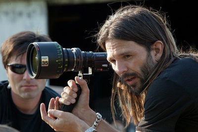 A man with long hair and a beard holds a professional camera lens while looking intently through the viewfinder.