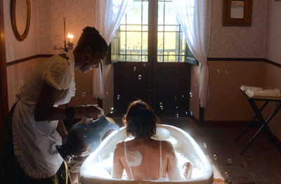 A maid in a white apron pours water from a pitcher into a bathtub for a woman bathing.