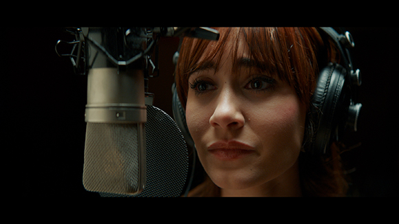 A woman with reddish-brown hair wearing headphones speaks into a large studio microphone.