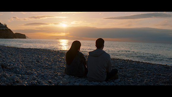 A man and woman sit on a rocky beach facing a sunset over the ocean horizon.