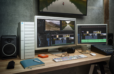 Dual monitor video editing setup with a large tower, studio speakers, keyboard, and editing software displayed.