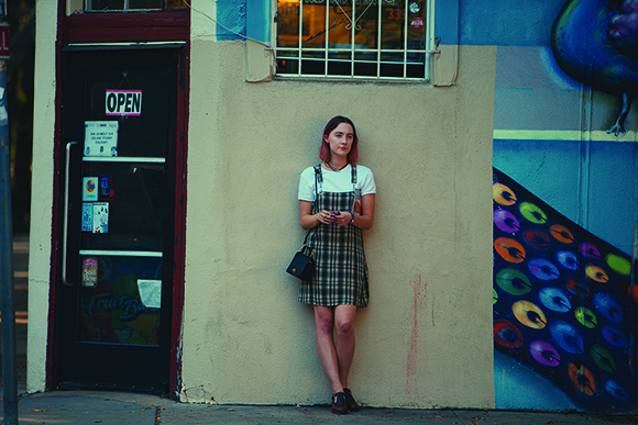 A woman in a white t-shirt and plaid jumper stands against a light-colored wall next to a colorful mural.