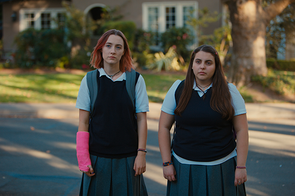 Two teenage girls in navy vests and grey skirts stand on a residential street.