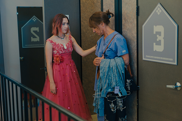 A woman in a pink formal dress talks to a woman in blue medical scrubs holding clothing.