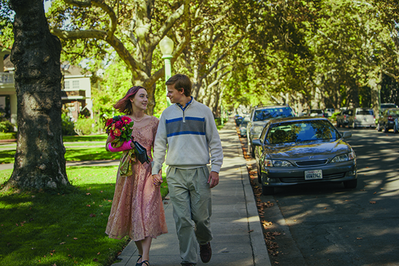 A young man and woman walk hand-in-hand along a sunny, tree-lined residential sidewalk.