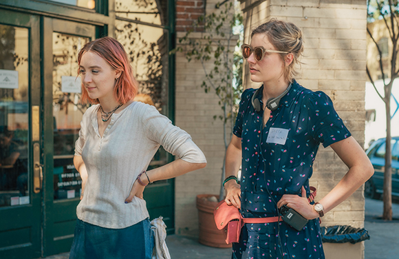 Two women stand outdoors near a green door; one wears a light shirt, the other a dark patterned dress and sunglasses.