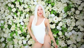 A woman with long blonde hair wearing a white bodysuit against a wall of white and green flowers.