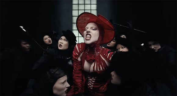 A woman in a red costume and large hat sings dramatically, surrounded by people in dark clothing.