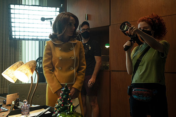 A woman in a yellow coat stands on a film set as a crew member in a green shirt points toward the distance.