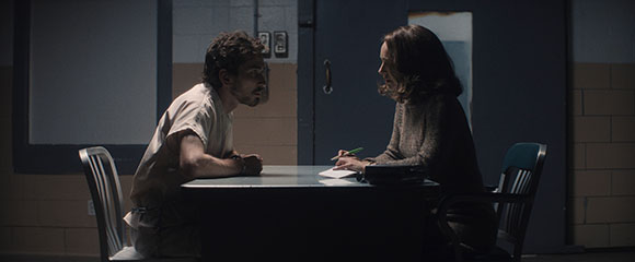 A man in a white shirt and a woman in a dark sweater sit at a table in a dark, sparse room.