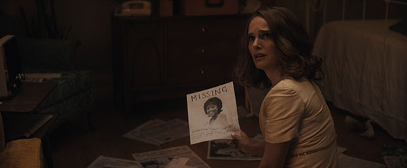 A woman in a light-colored dress holds a flyer with a person's photo in a dark room.