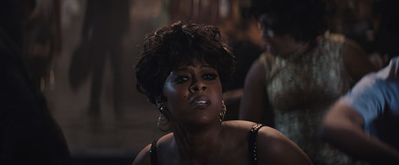 A woman with dark curly hair and hoop earrings looks upward in a dark, crowded room.