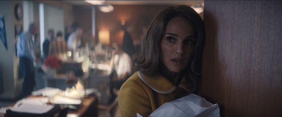 A woman with wavy brown hair peeks cautiously from behind a wooden wall in a crowded, dimly lit room.