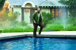 A man in a green jacket and brown pants stands at the edge of a blue swimming pool near a small house.