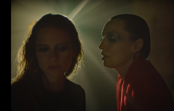 A person with dark hair and makeup looks toward another person under a dramatic beam of light.
