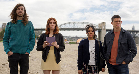 Four people stand on a sandy beach with a large metal bridge under a cloudy sky.