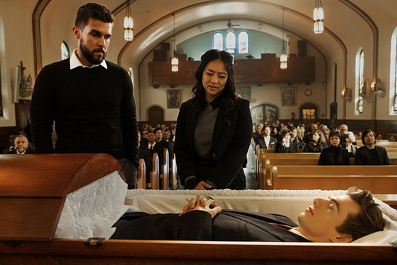 A man and a woman stand solemnly beside an open wooden casket in a church.