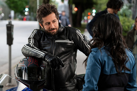 A man in a black leather jacket leans on a motorcycle while talking to a woman with long dark hair.