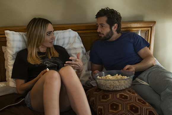 A man and woman sit on a bed together, sharing a large bowl of popcorn.