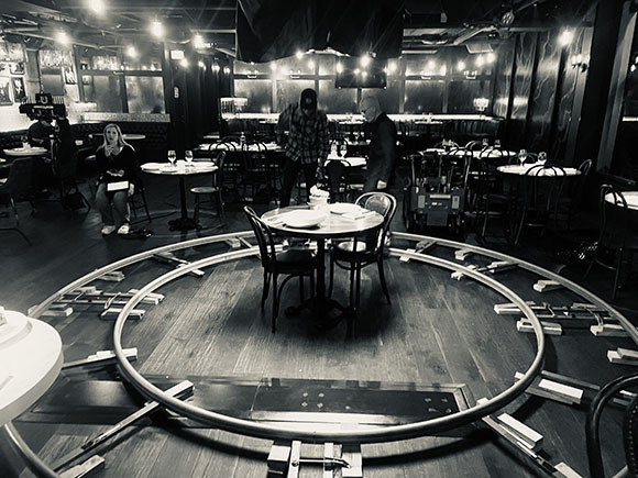 Black and white wide shot of a film crew setting up equipment in a restaurant with circular tracks on the floor.