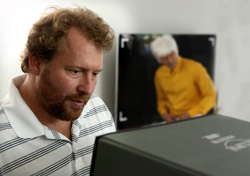 A man with curly hair and a beard looks down at a black box in a bright room.