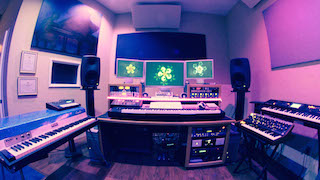 A music studio featuring three computer monitors, studio monitors, and several electronic keyboards.